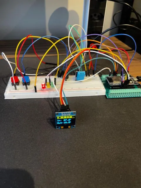 A protoboard do Atmos sobre a mesa: o ESP32 à direita, o sensor de temperatura no centro, botões e LEDs à esquerda, dezenas de jumpers coloridos, e o display OLED à frente mostrando as temperaturas de dentro e de fora.